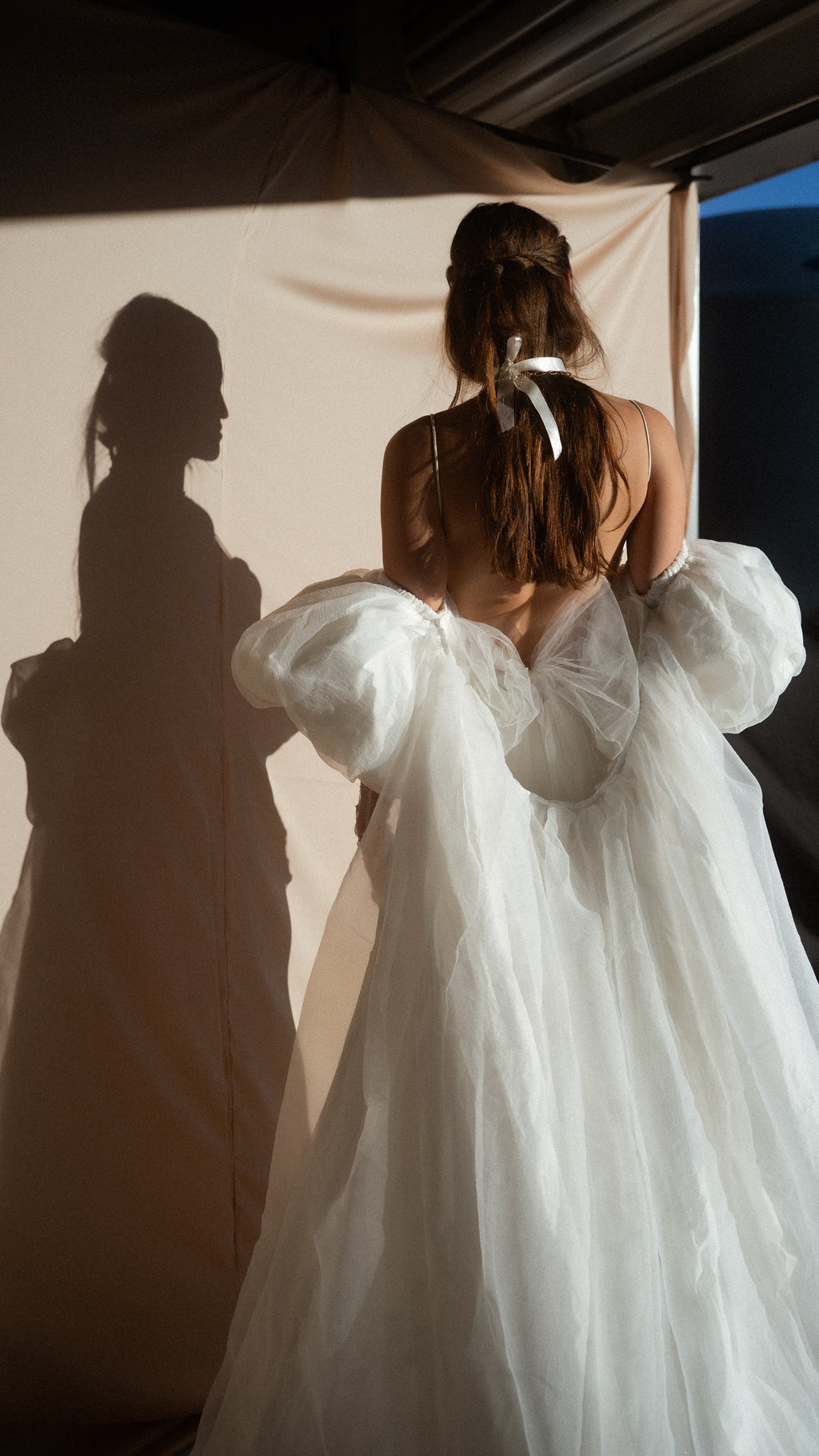 Three-quarter back view of an ivory bridal cape, chiffon puff sleeves framing a bare back, shadow cast on a draped wall, Aura Cape