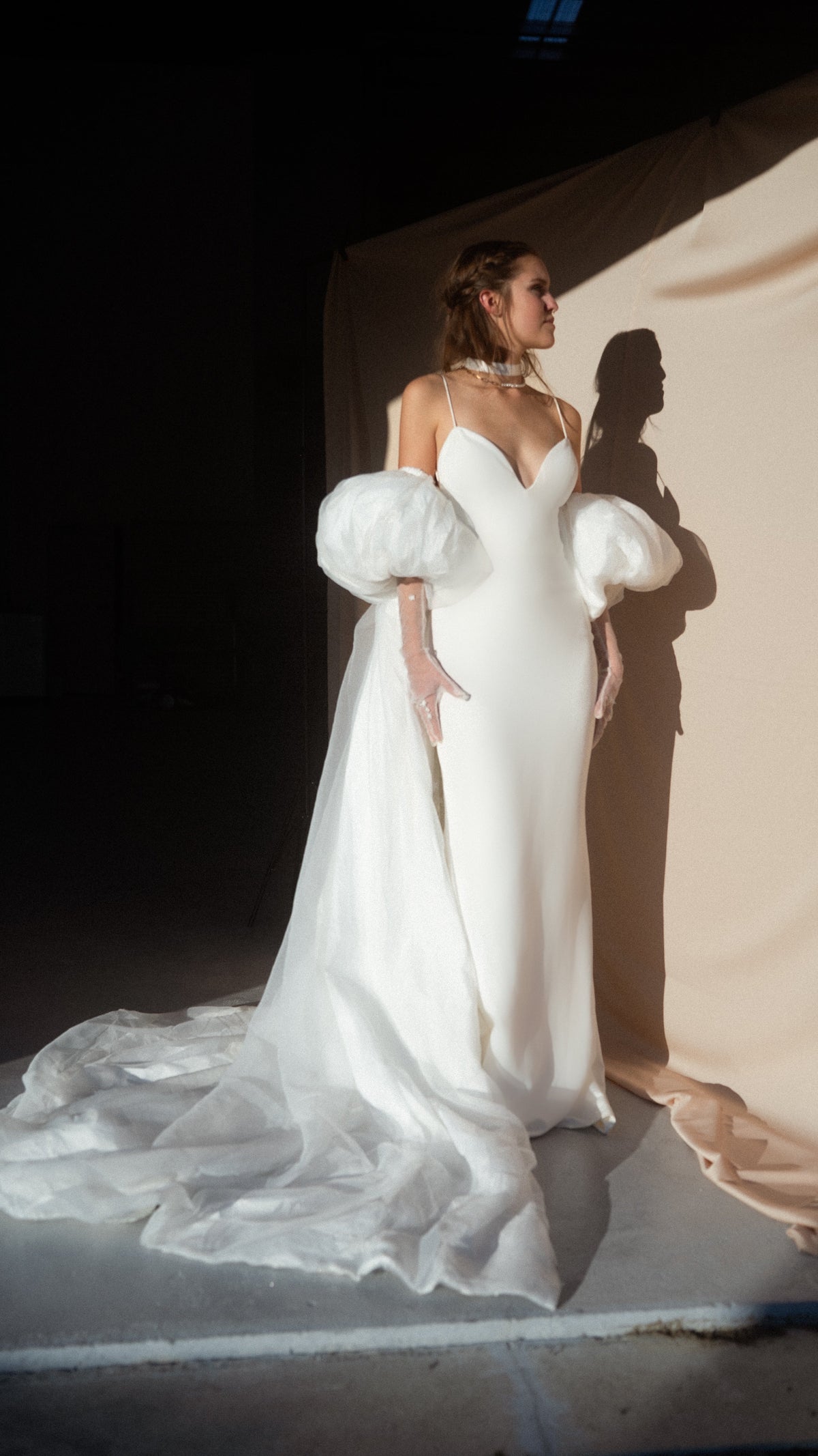 Ivory bridal cape with puff sleeves and long chiffon train, bride in profile against a draped backdrop in dramatic light, Aura Cape