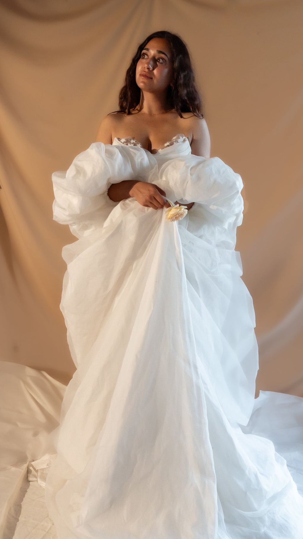 Bride holding a flower in an ivory bridal cape with crinkled chiffon puff sleeves gathered at the front, warm studio backdrop, Aura Cape