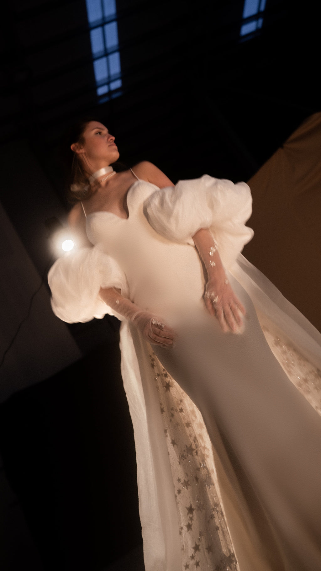 Low-angle editorial shot of an ivory bridal cape with puff sleeves and a starry lining, bride leaning back under a spotlight, Aura Cape