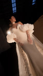 Low-angle editorial shot of an ivory bridal cape with puff sleeves and a starry lining, bride leaning back under a spotlight, Aura Cape