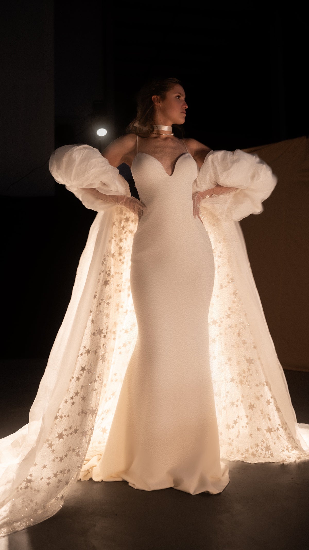 Celestial bridal cape with stars glittering along the inner train, puff sleeves and an ivory slip gown, backlit studio, Aura Cape