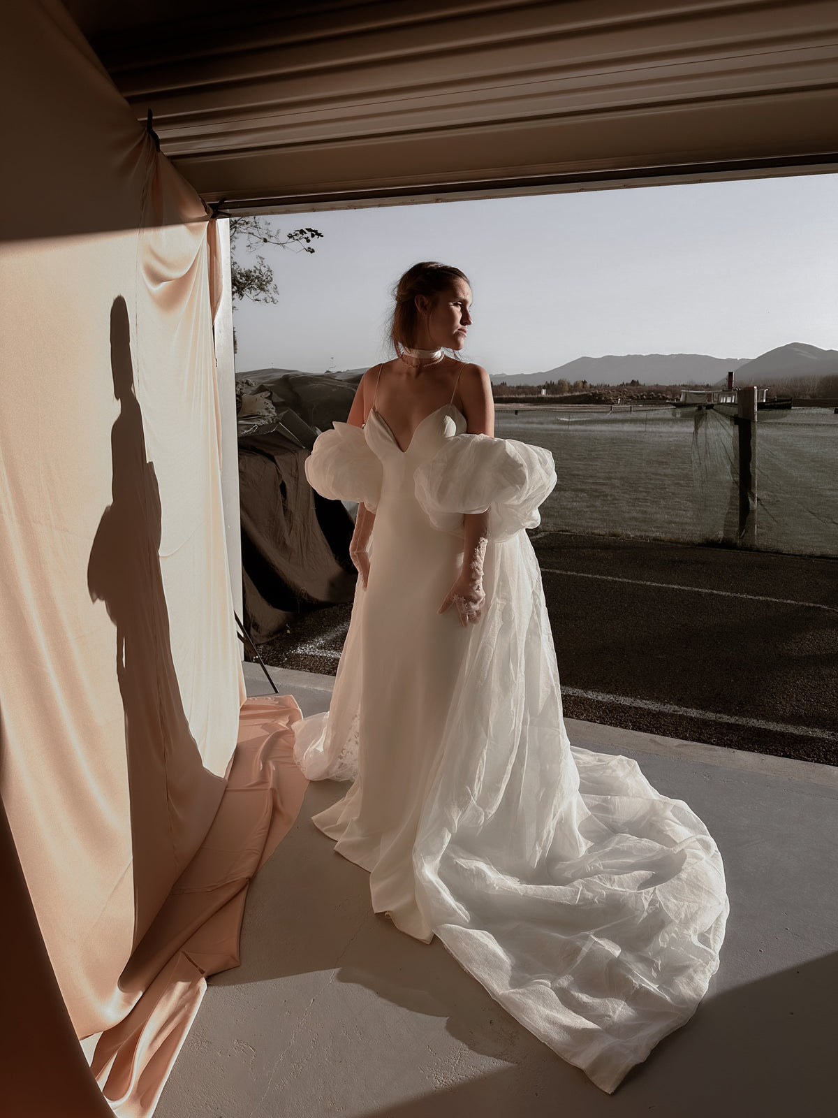Bride standing in an open doorway with a lake view, ivory bridal cape train trailing over the floor, puff sleeves, Aura Cape
