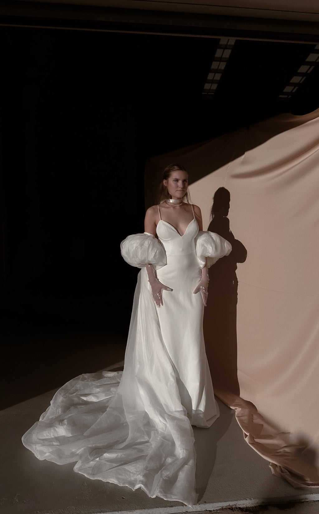 Ivory bridal cape with puff sleeves over a slip gown, chiffon train curving across a studio floor in low light, Aura Cape