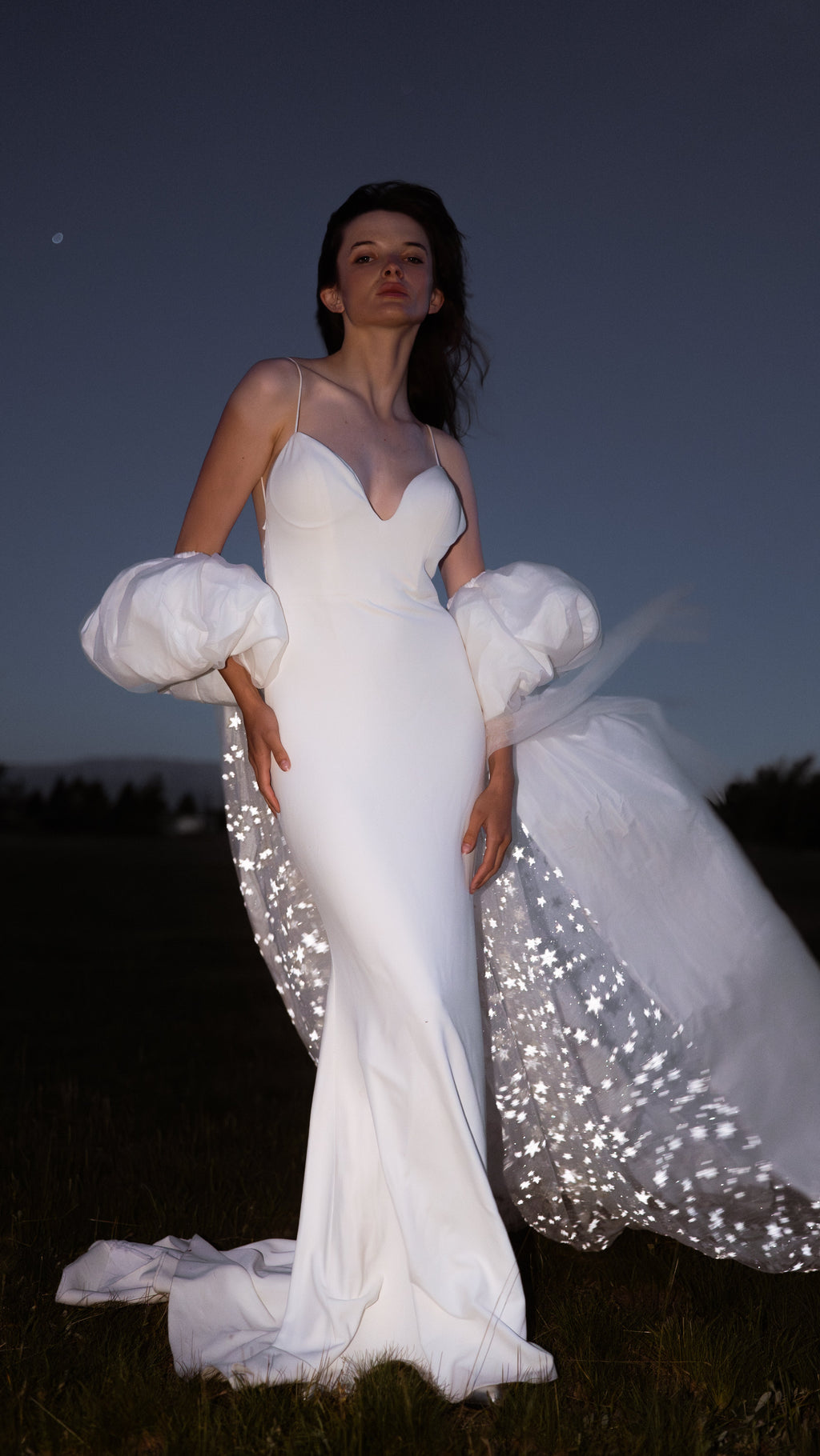 Bride in a field after dark, ivory bridal cape with puff sleeves and a starry train blowing out to one side, Aura Cape