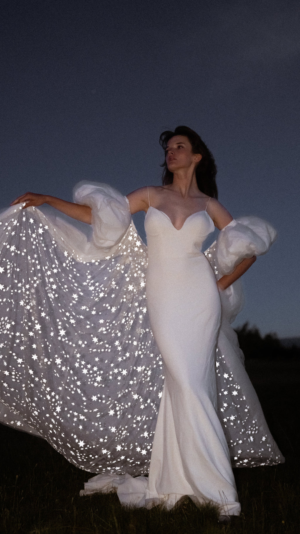 Bride holding one side of a bridal cape open to reveal a sky of glittering stars inside the train, night field, Aura Cape