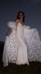 Ivory bridal cape held open with both hands, star-covered lining shining around a slip gown, low-angle night portrait, Aura Cape