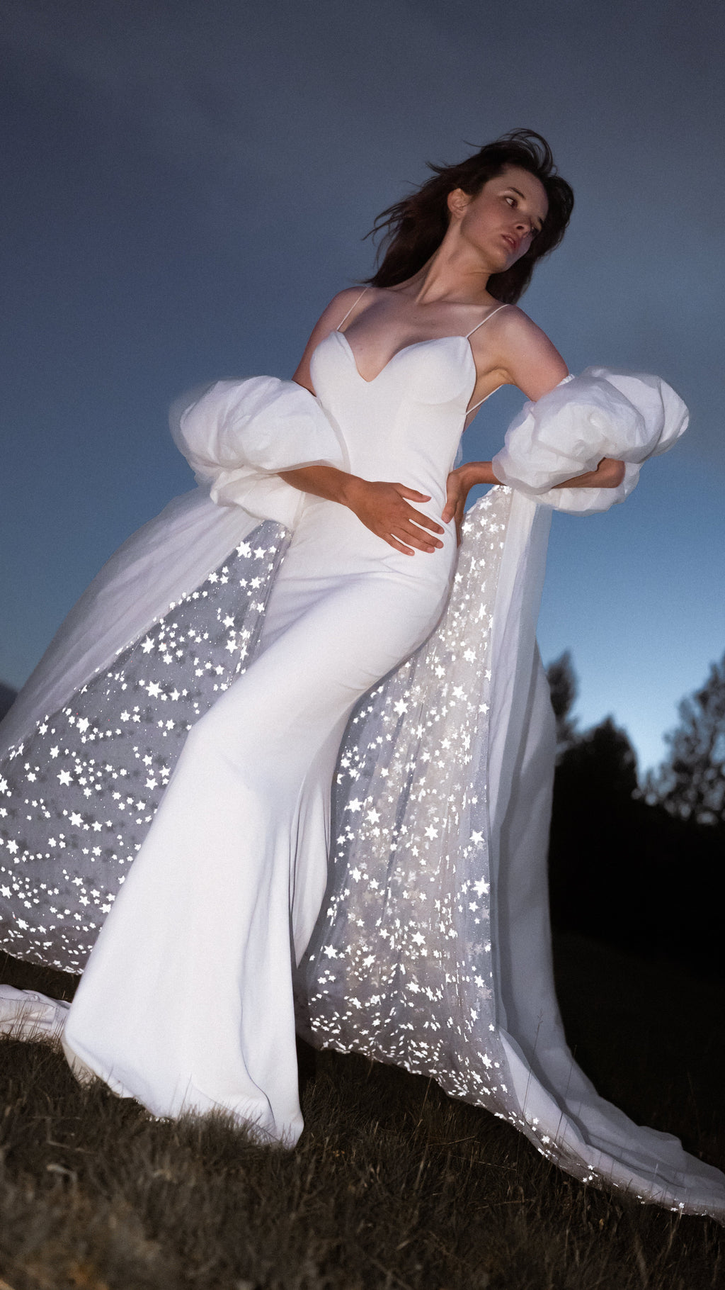 Bride with hands on her waist in an ivory bridal cape, puff sleeves and a star-lined train spread behind her at dusk, Aura Cape