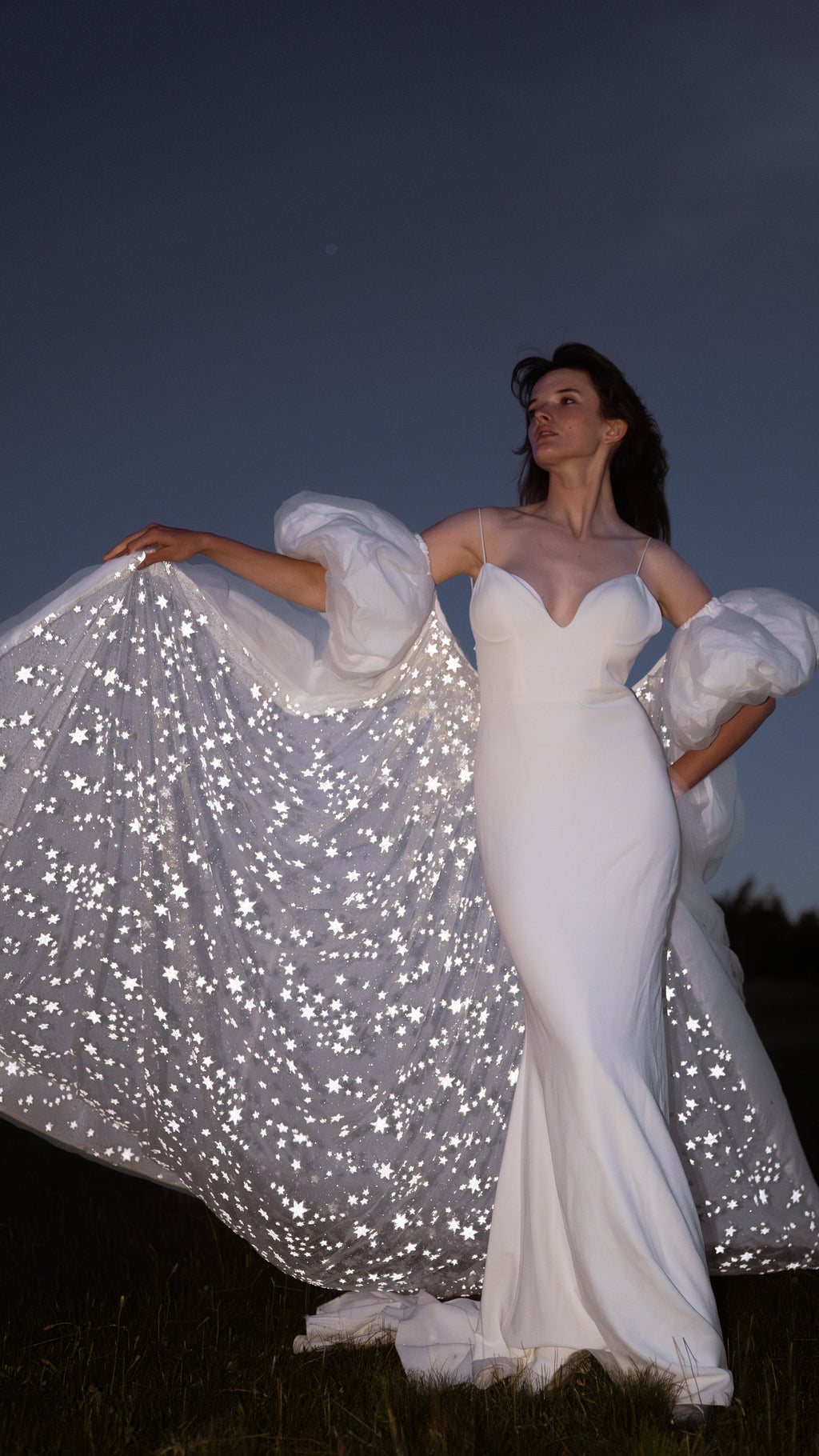 Bridal cape lifted wide by one hand like a wing, stars glittering across the train against a night sky, Aura Cape