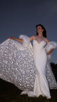 Bridal cape lifted wide by one hand like a wing, stars glittering across the train against a night sky, Aura Cape