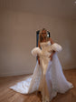 Wide room view of a bride in an ivory bridal cape with puff sleeves over a satin corset gown, train fanned behind, Aura Cape