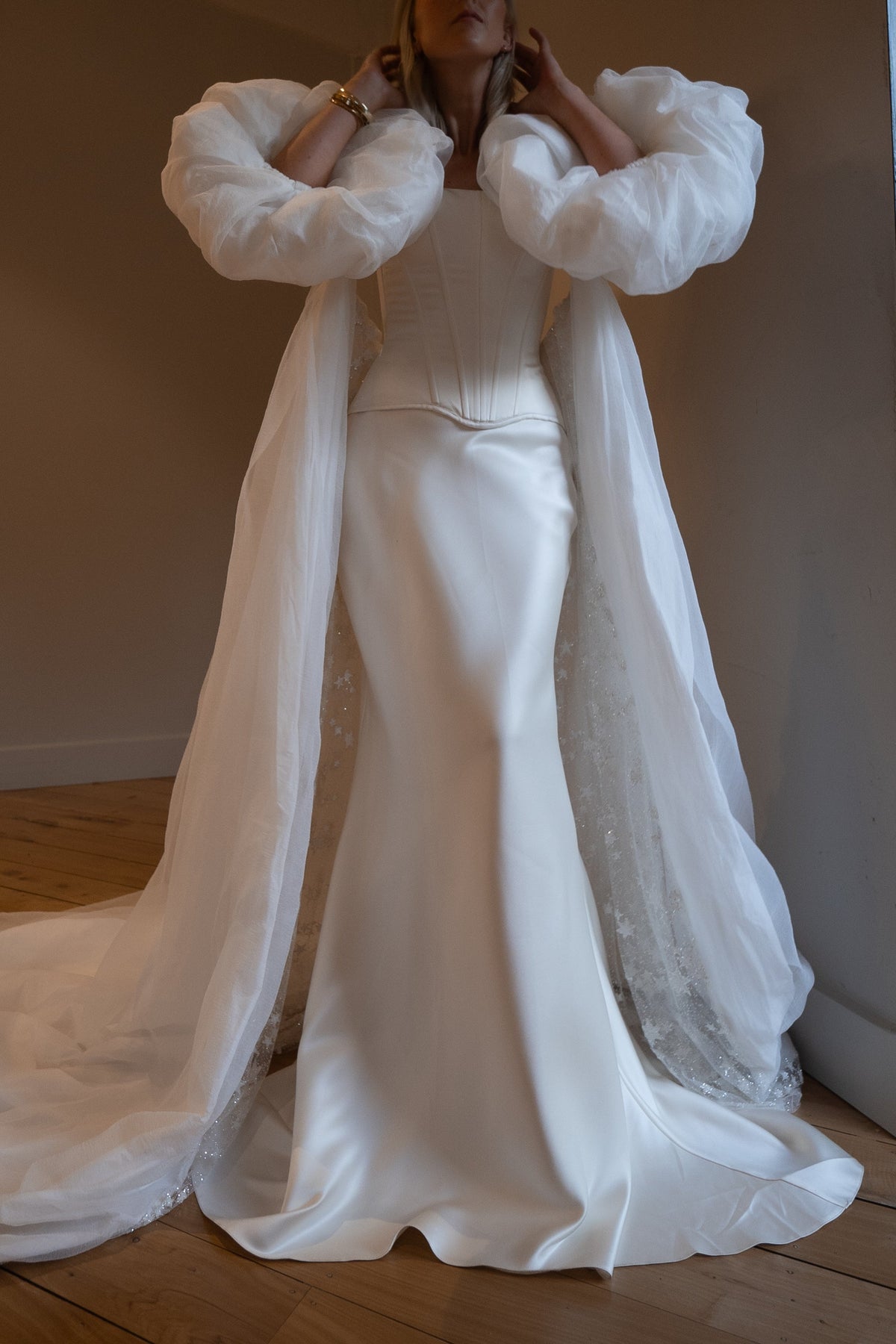 Bride with hands raised to her hair, ivory bridal cape with oversized puff sleeves and a train over a satin corset gown, Aura Cape