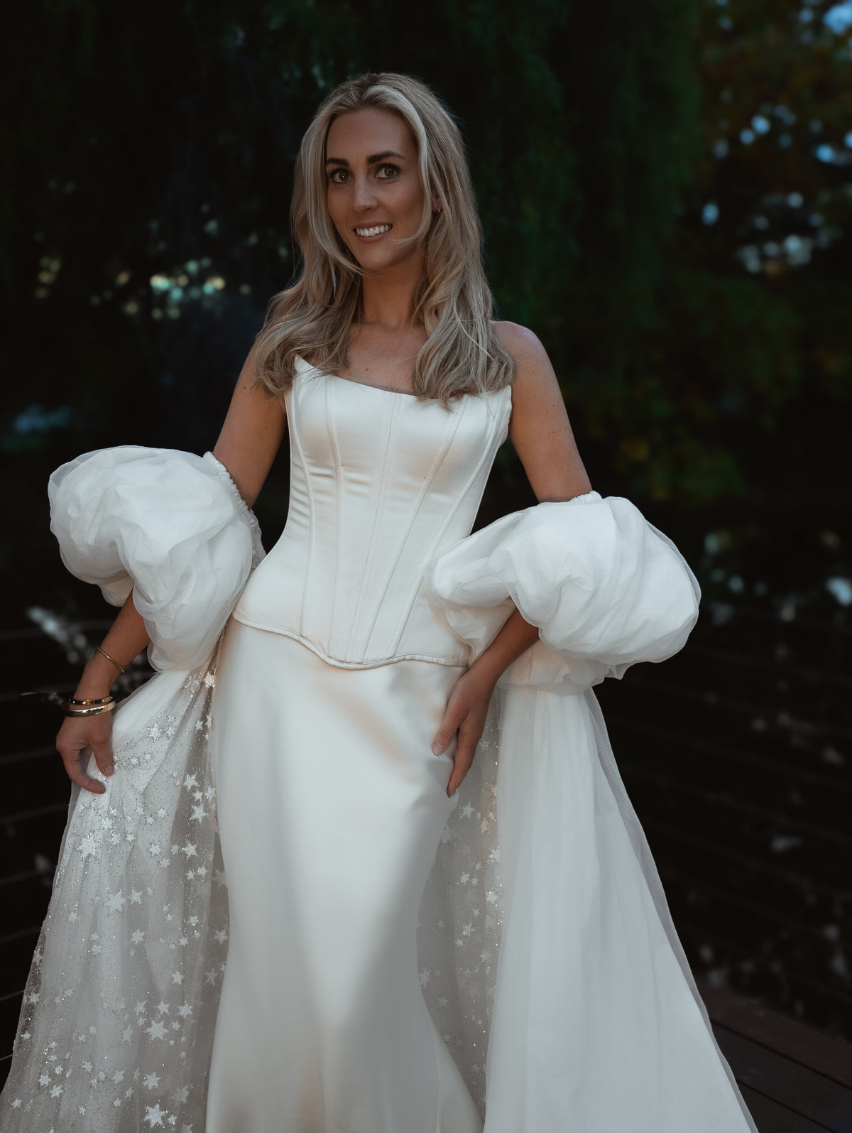 Smiling bride in an ivory bridal cape with chiffon puff sleeves and a star-scattered train, garden at dusk, Aura Cape