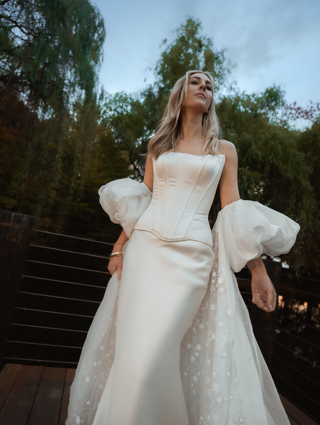 Low-angle view of an ivory bridal cape with puff sleeves and star tulle train over a satin corset gown, trees behind, Aura Cape