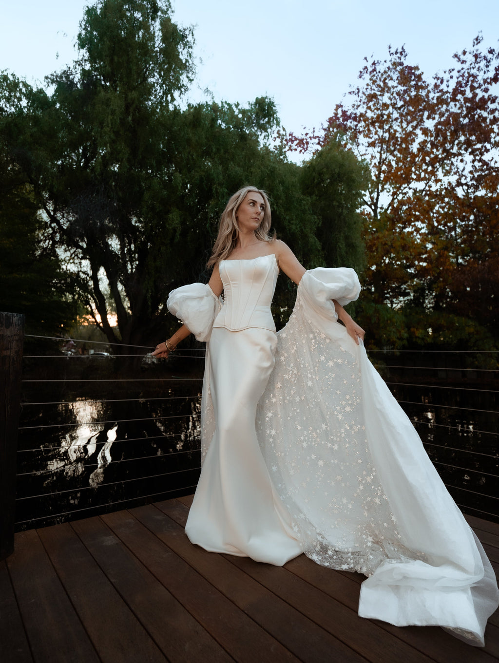 Bride on a deck by a pond, ivory bridal cape with puff sleeves and a long star-dotted train stretched across the boards, Aura Cape