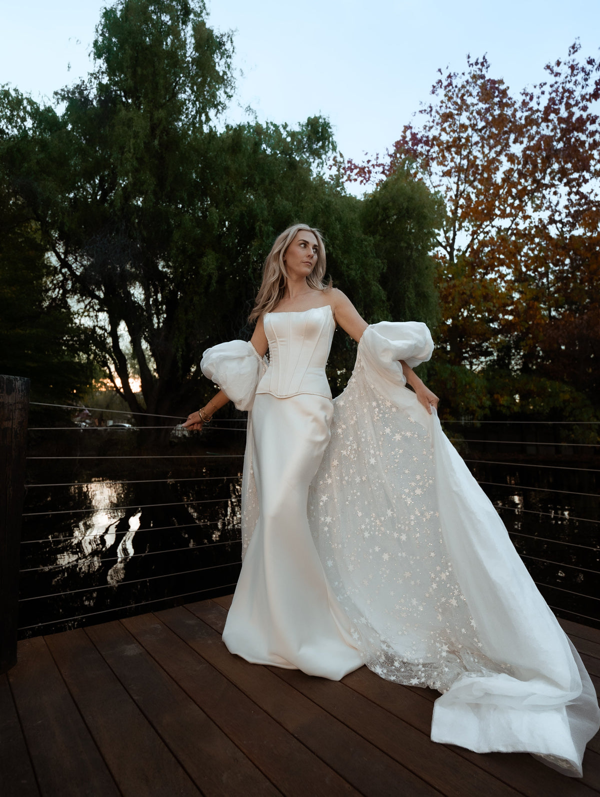 Bride on a deck by a pond, ivory bridal cape with puff sleeves and a long star-dotted train stretched across the boards, Aura Cape
