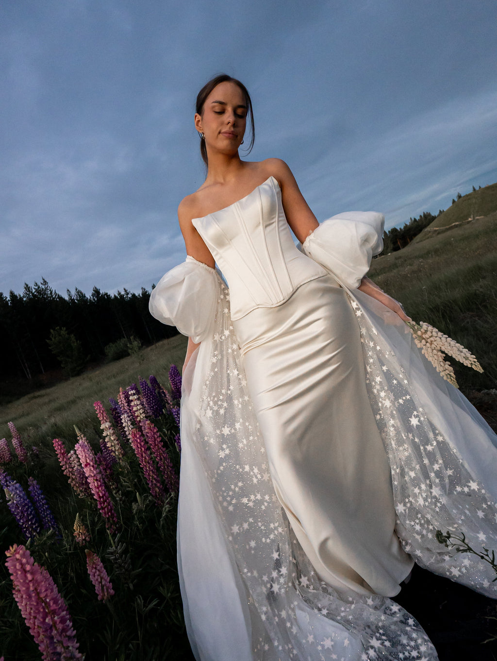 Bridal cape with a starry tulle train worn over a satin corset gown, bride in a field of pink and purple lupines, Aura Cape