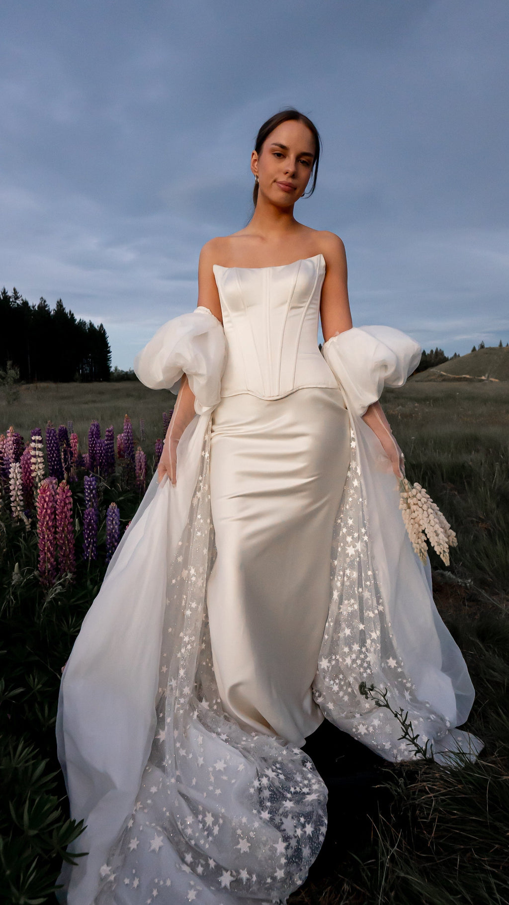Bridal cape with puff sleeves and a starry inner train worn over a satin corset gown in a wildflower field of lupines, Aura Cape