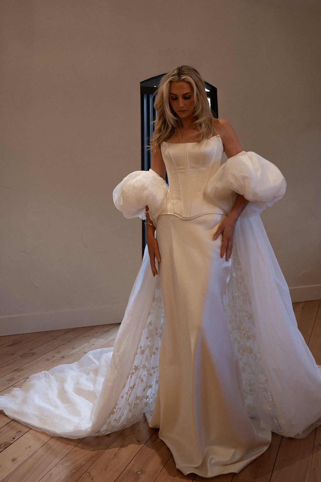 Ivory bridal cape with puff sleeves over a satin corset gown, train spread on a wooden floor in a plain studio room, Aura Cape