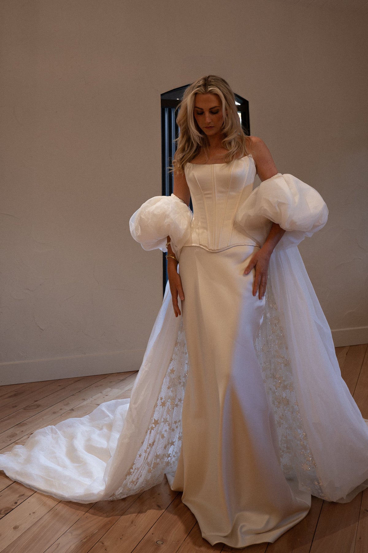 Ivory bridal cape with puff sleeves over a satin corset gown, train spread on a wooden floor in a plain studio room, Aura Cape