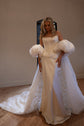 Ivory bridal cape with puff sleeves over a satin corset gown, train spread on a wooden floor in a plain studio room, Aura Cape