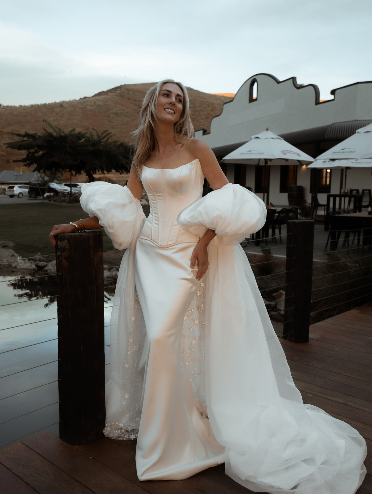 Bride leaning on a deck railing in an ivory bridal cape with chiffon puff sleeves and a long train, outdoor lodge at dusk, Aura Cape