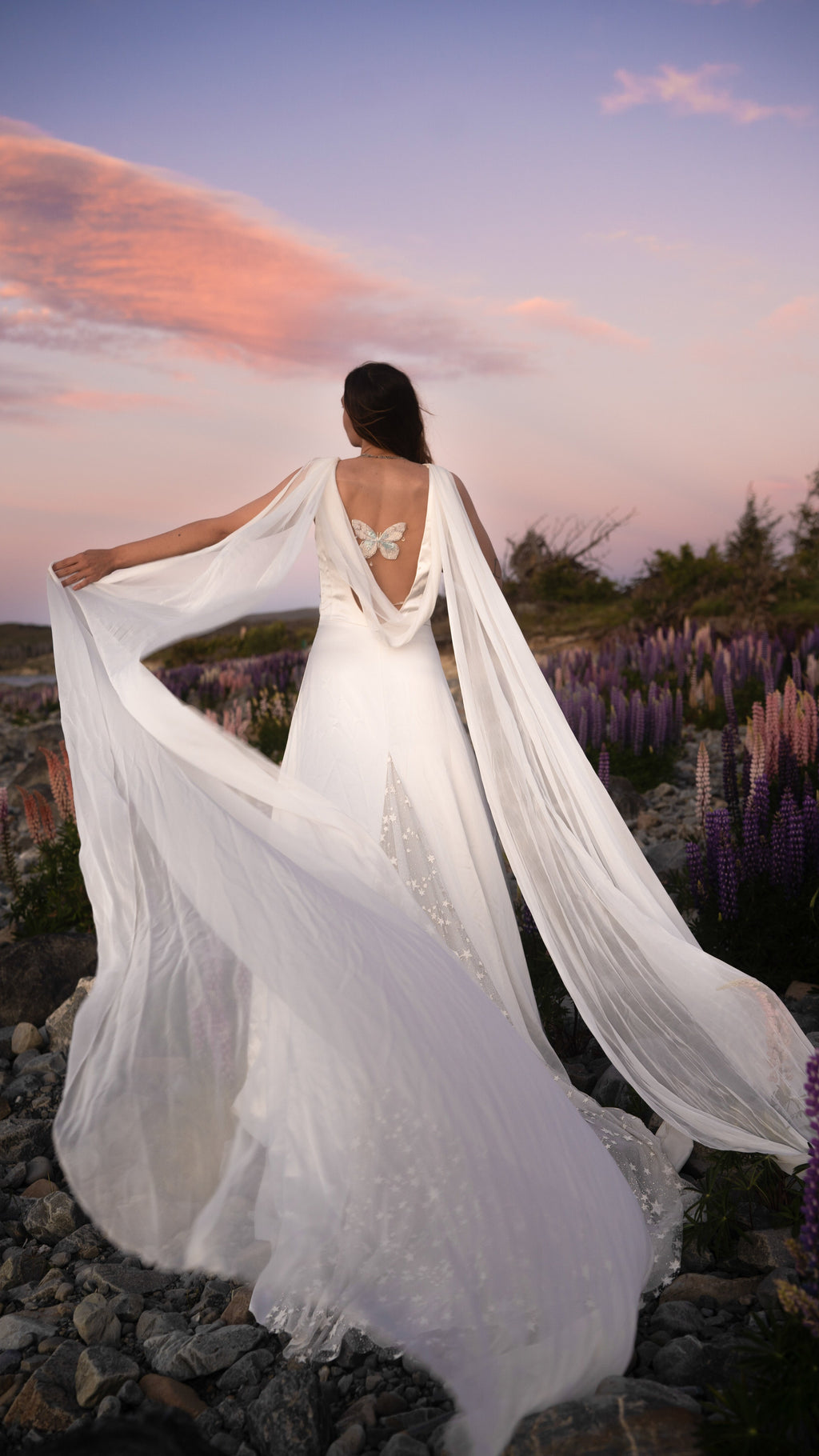 Open back satin wedding dress with silk chiffon wings and a star-scattered train, bride in a lupine field at sunset. Aurora by Ephemeris