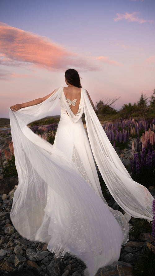Open back satin wedding dress with silk chiffon wings and a star-scattered train, bride in a lupine field at sunset. Aurora by Ephemeris