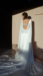 Full-length back view of a satin wedding dress with open cowl back, chiffon wings pooling on the floor in warm light, Aurora
