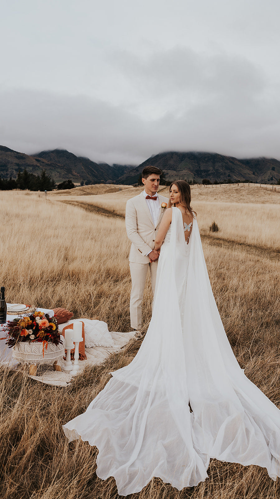 Elopement picnic in a golden grass field, bride in a satin wedding dress with long chiffon wings beside the groom, Aurora