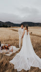 Elopement picnic in a golden grass field, bride in a satin wedding dress with long chiffon wings beside the groom, Aurora
