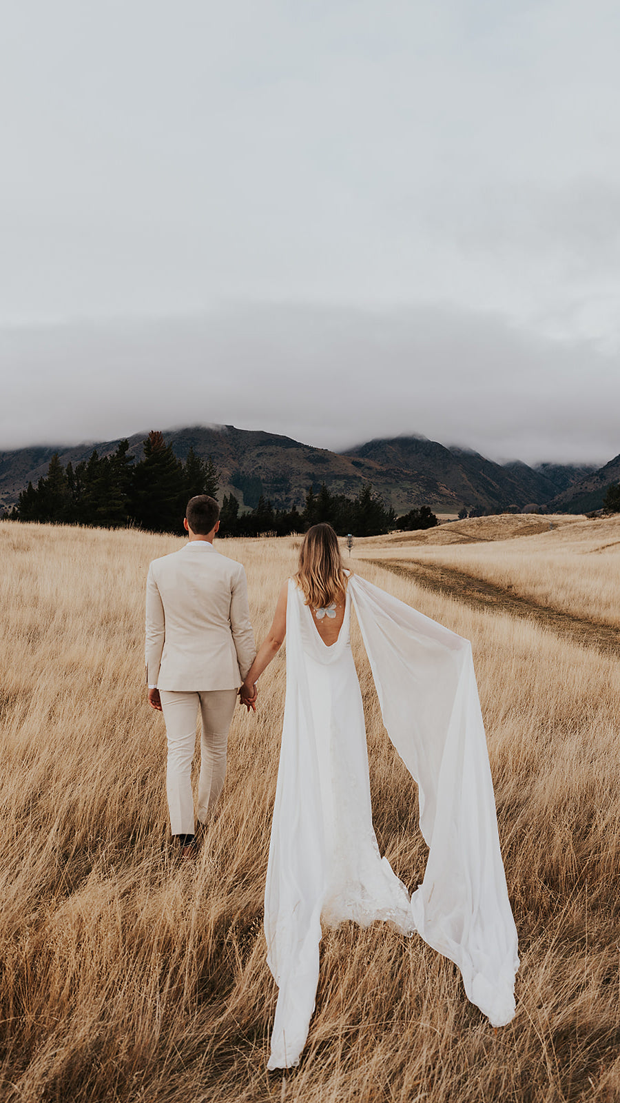 Couple walking hand in hand through tall grass, chiffon wings of a satin wedding dress trailing behind the bride, Aurora