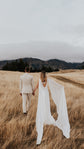 Couple walking hand in hand through tall grass, chiffon wings of a satin wedding dress trailing behind the bride, Aurora