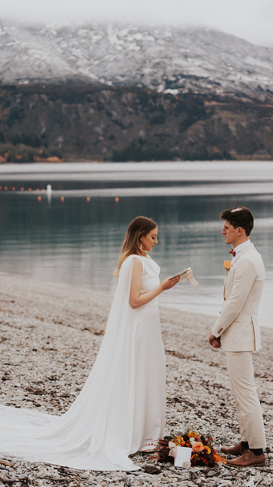 Bride reading vows on a lakeside shore in a satin wedding dress with chiffon wings, mountains behind, Aurora