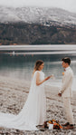 Bride reading vows on a lakeside shore in a satin wedding dress with chiffon wings, mountains behind, Aurora
