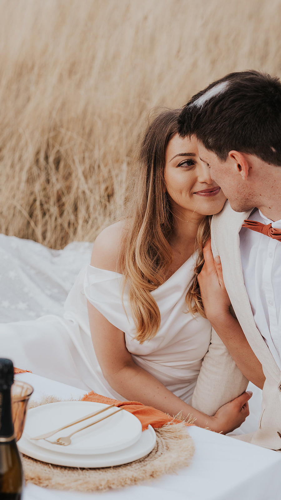Close-up of a smiling bride in a satin cowl neck wedding dress leaning on the groom at a picnic table, Aurora