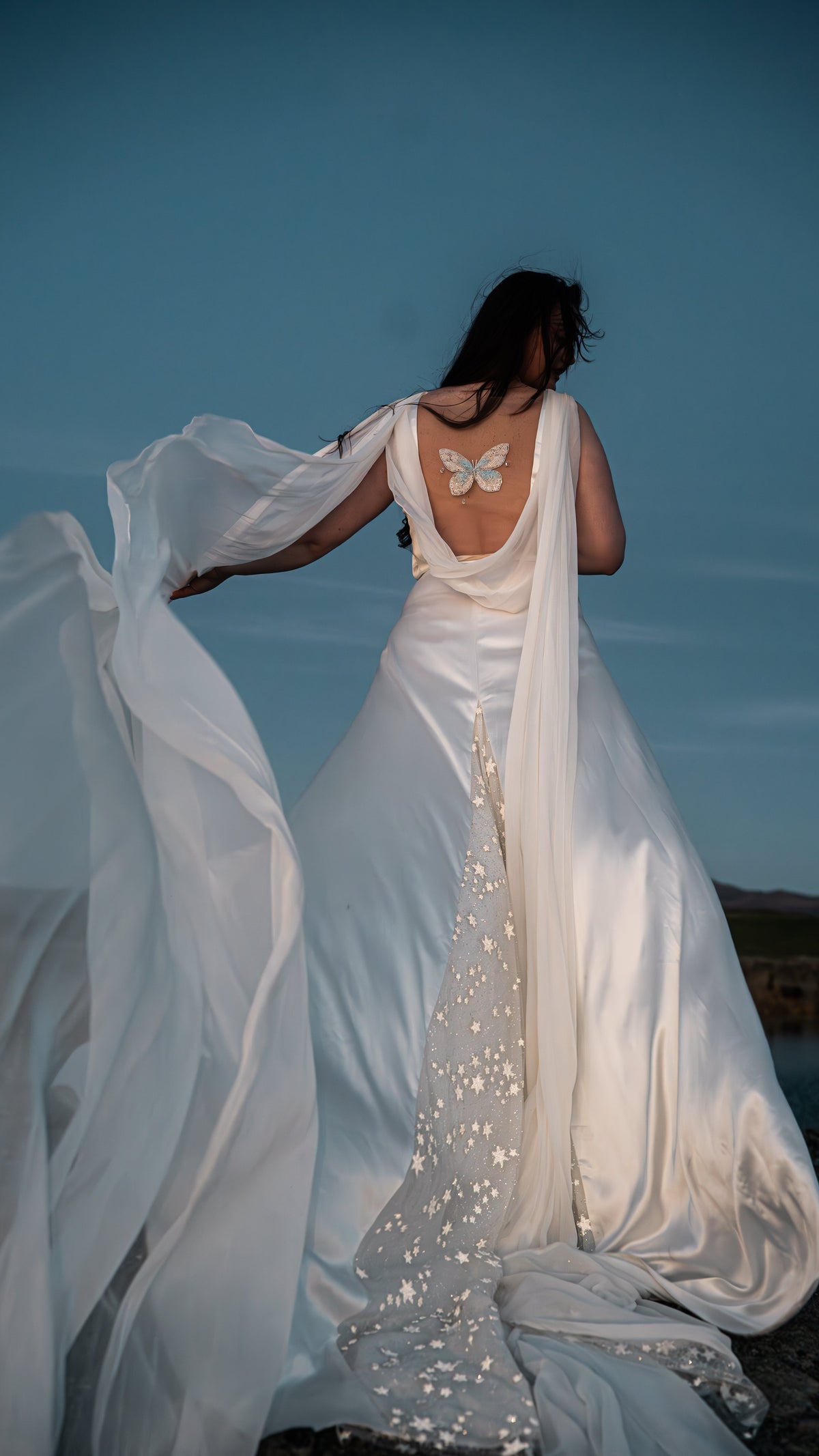 Wind lifting a chiffon wing off a satin open back wedding dress, butterfly embroidery and star train at blue hour, Aurora
