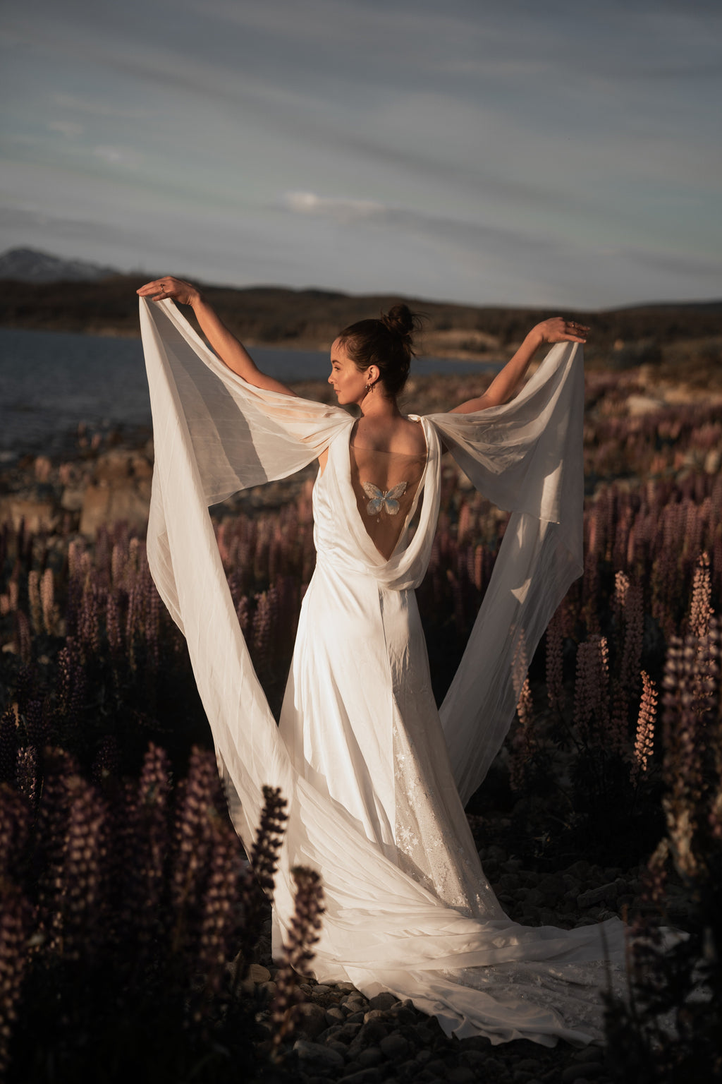Bride raising silk chiffon wings overhead in a wildflower field, open back satin wedding dress in evening light, Aurora