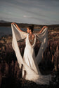 Bride raising silk chiffon wings overhead in a wildflower field, open back satin wedding dress in evening light, Aurora