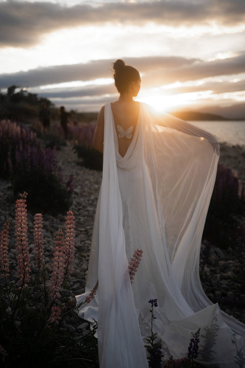 Back view of a satin wedding dress with cowl open back and butterfly embroidery, chiffon wings held out against the setting sun, Aurora