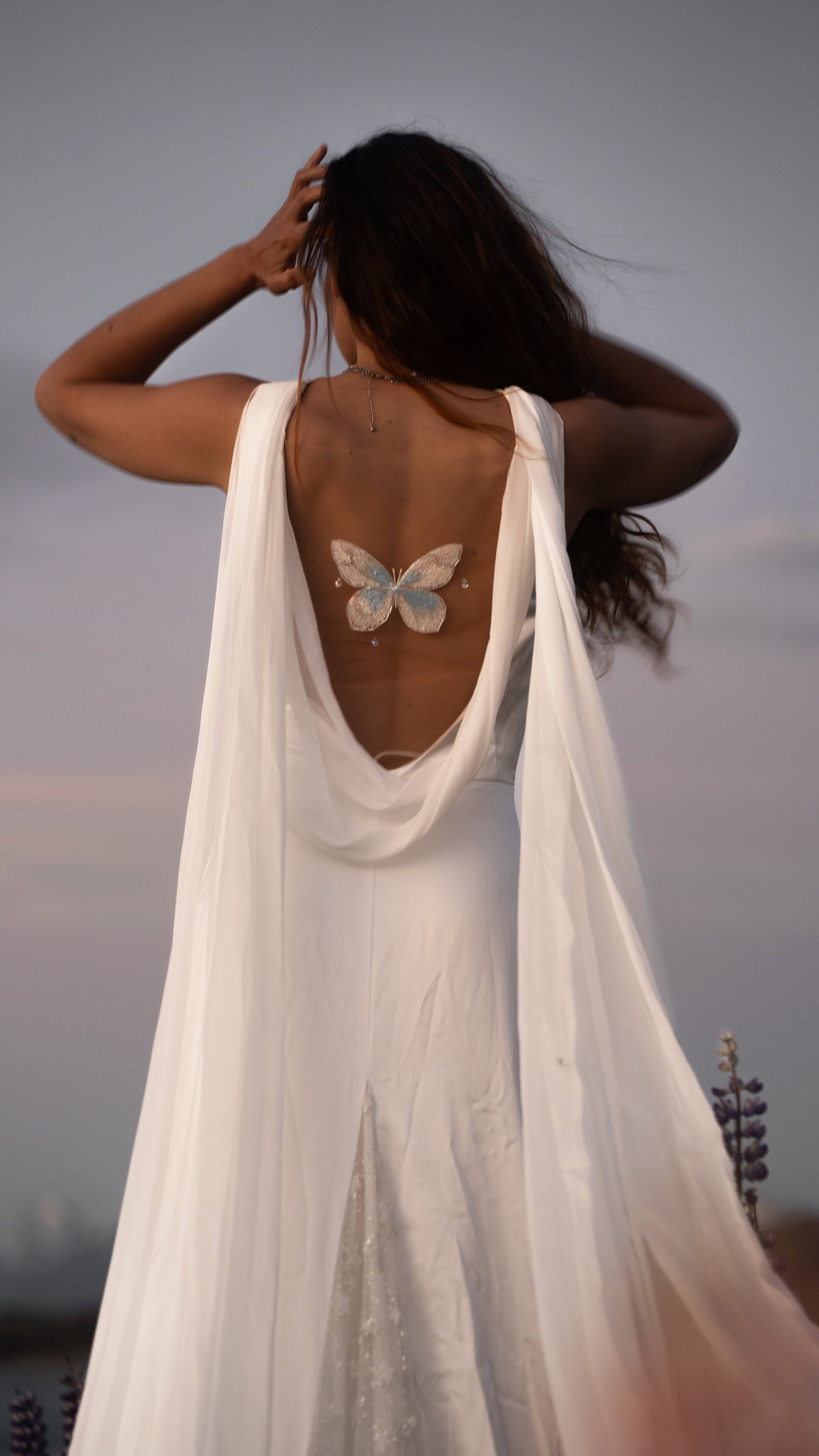 Close-up of the draped open back of a satin wedding dress with hand embroidered butterfly and chiffon wings, bride touching her hair, Aurora