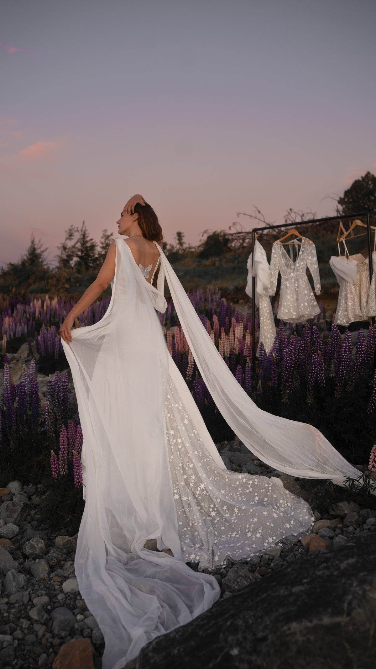 Bride turning in a satin wedding dress with long chiffon wings and a star-dotted train, lupines and a dress rail behind, Aurora
