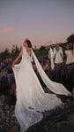 Bride turning in a satin wedding dress with long chiffon wings and a star-dotted train, lupines and a dress rail behind, Aurora