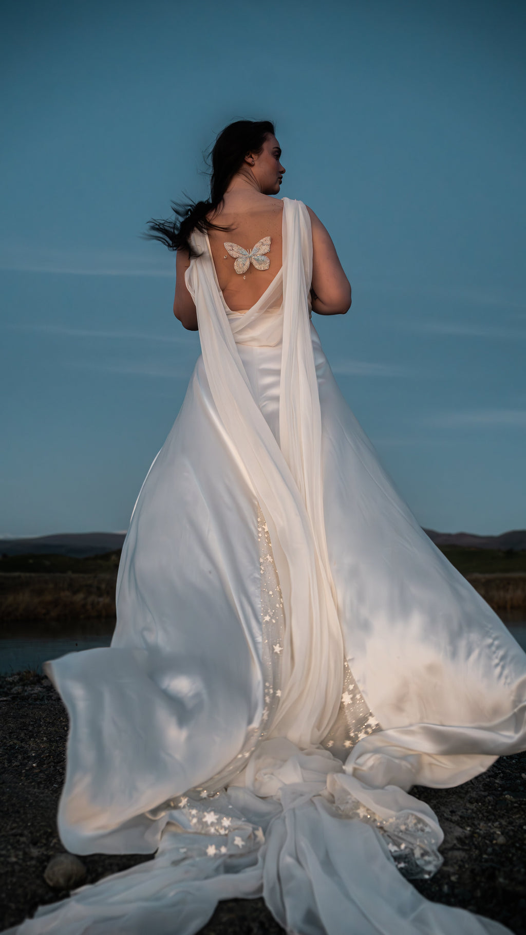 Satin wedding dress with long chiffon wings and star-scattered train spread over stones, bride looking back at blue hour, Aurora
