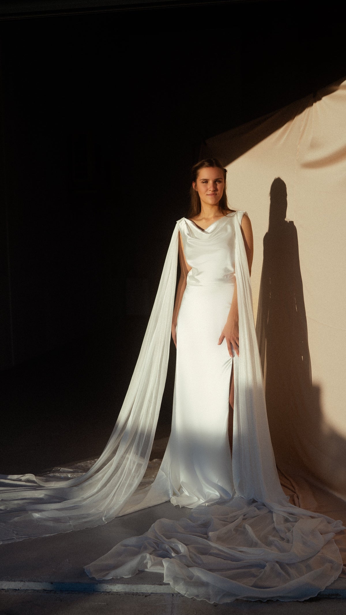 Front view of a satin wedding dress with cowl neckline and floor-length chiffon wings, bride standing in sharp studio light, Aurora