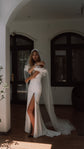 Sample wedding dress in stretch double crepe with tulle ruffle shoulders and a high slit, bride in a long veil by arched windows. Celeste Sample by Ephemeris