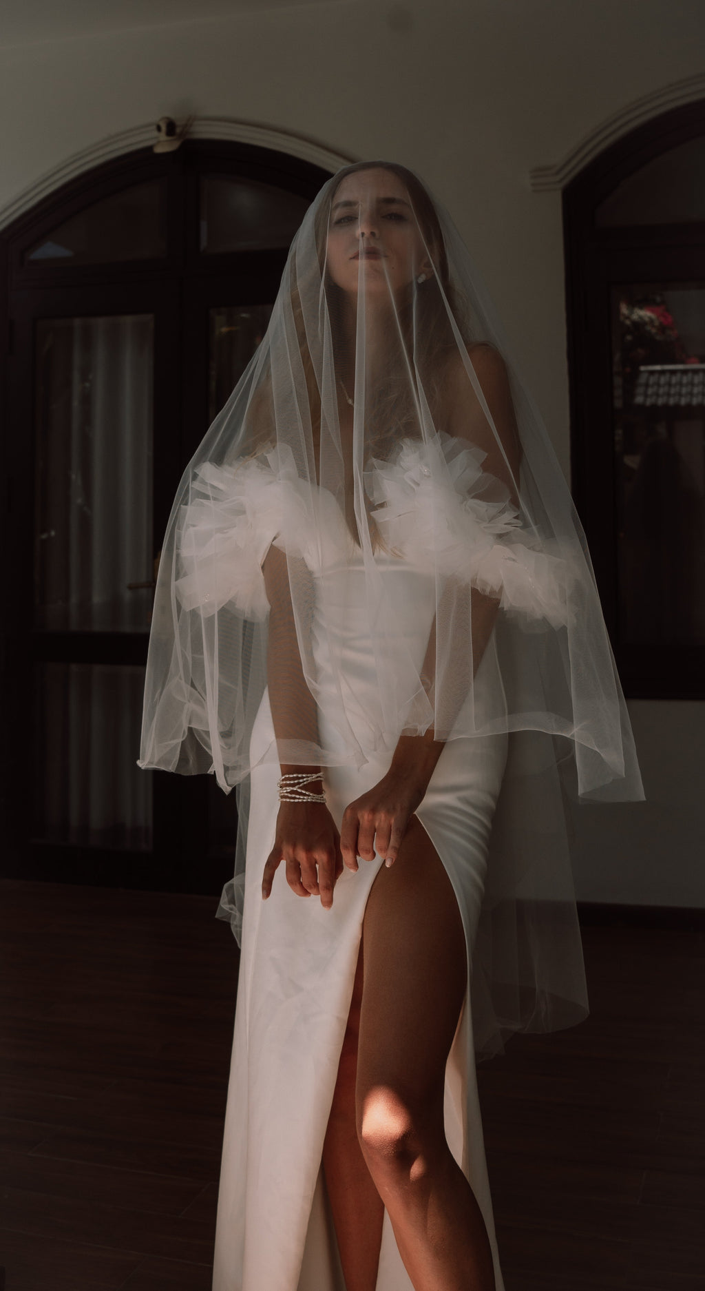 Off-shoulder crepe sample wedding dress with tulle ruffles, bride under a short sheer blusher veil in a dim room, Celeste Sample
