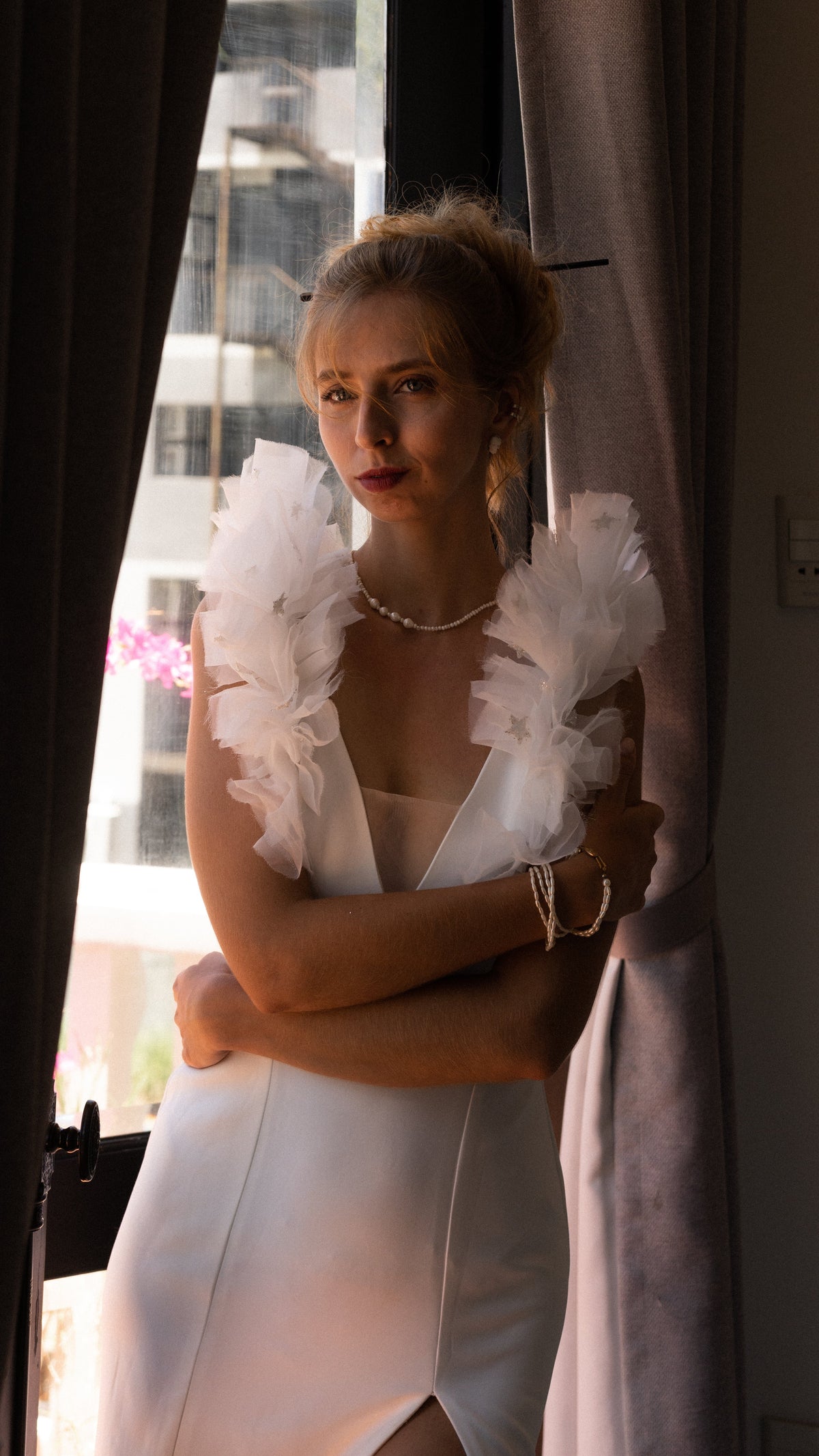 Portrait by a curtain in a V-neck crepe sample wedding dress, star-dotted tulle ruffles at the shoulders, arms crossed, Celeste Sample
