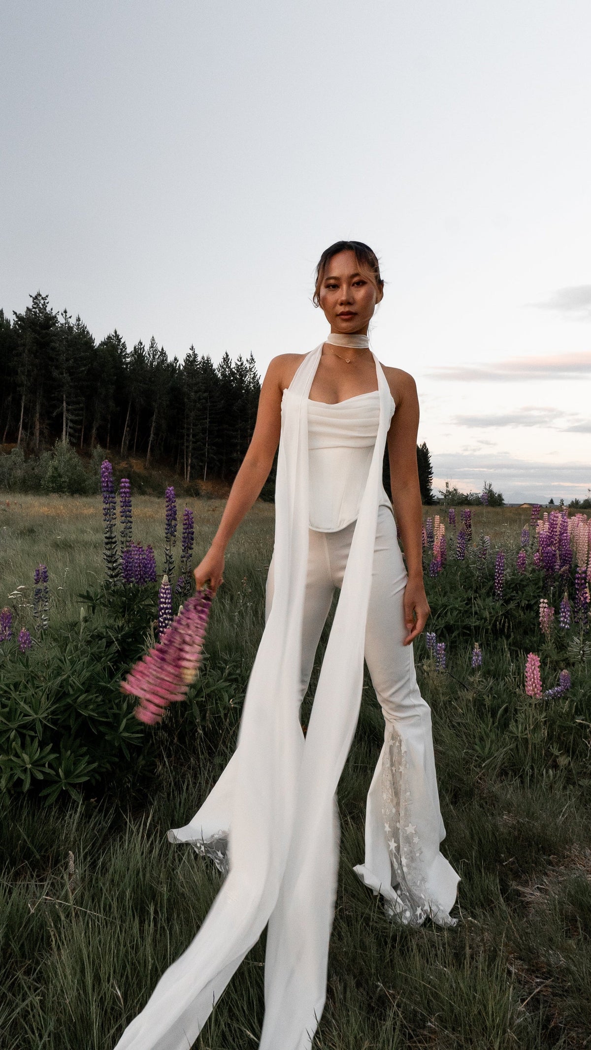 Long chiffon bridal scarf worn as a neck tie over a strapless top and flared trousers, lupines in hand at dusk, Celeste Scarf