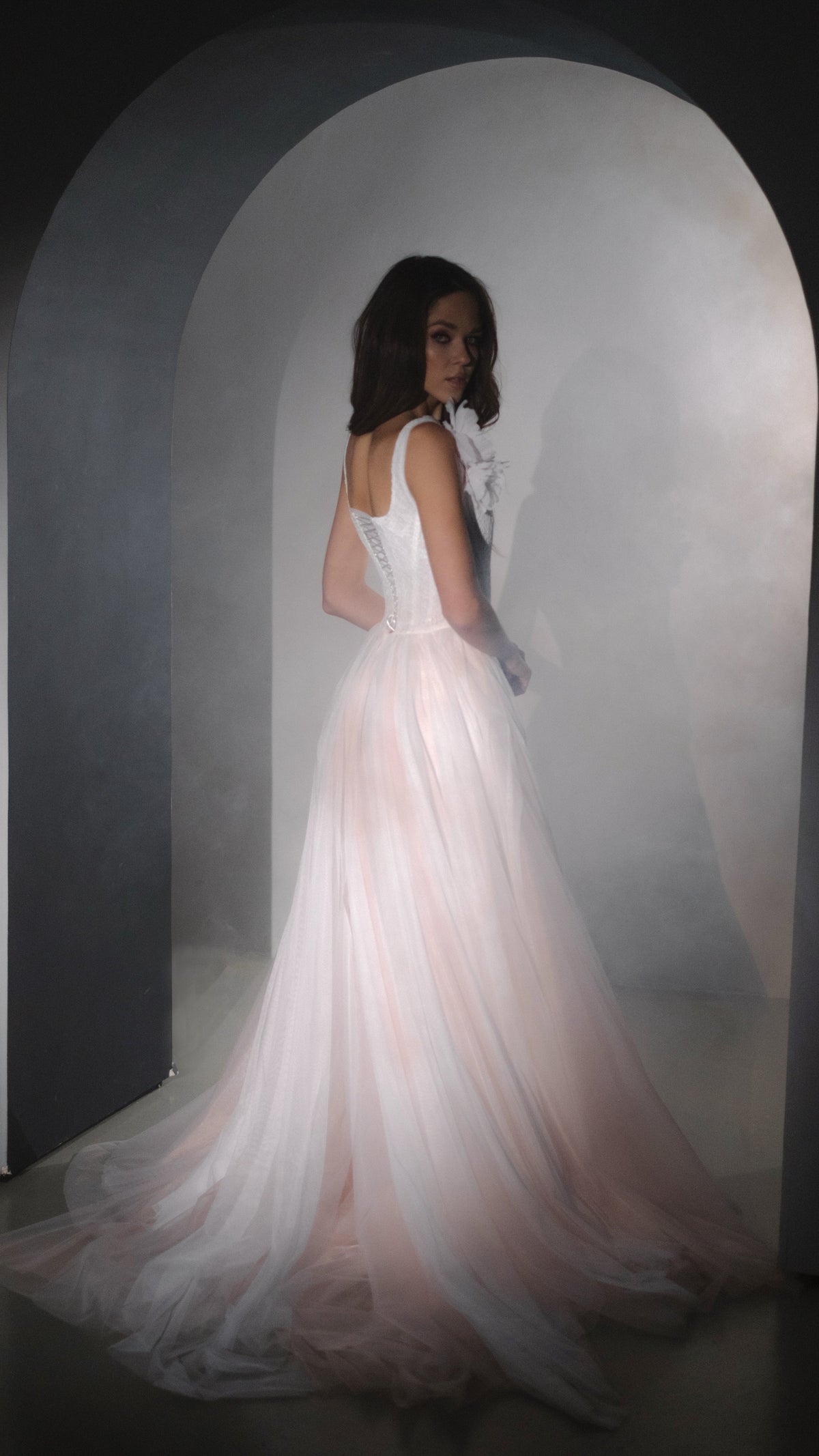 Three-quarter back view of a blush pink wedding dress with laced corset and long tulle train in an arched alcove, Chrysanthemum